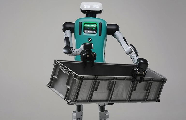 Digit bipedal humanoid robot standing in an industrial warehouse fulfillment aisle carrying automated logistics totes.