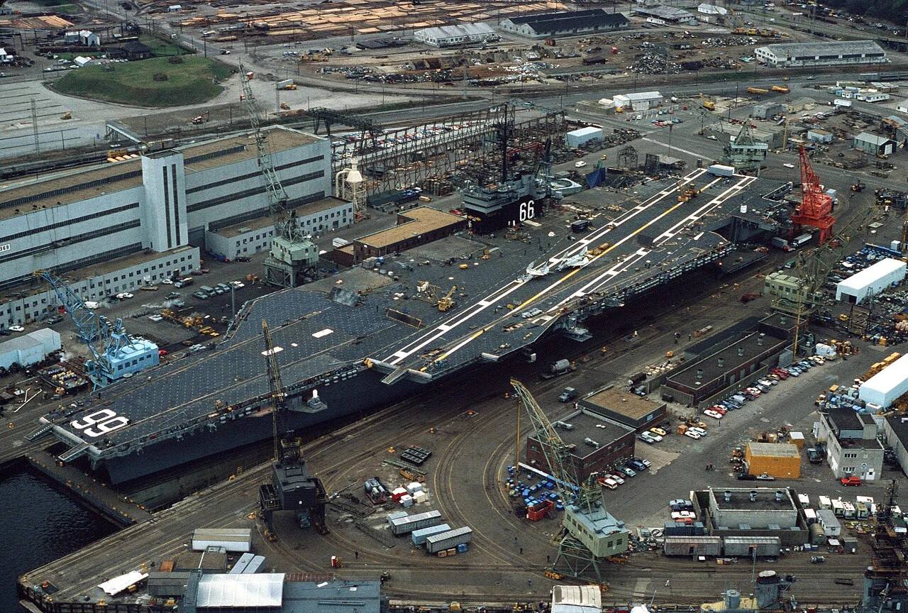 Naval warship inside shipyard dry dock facility undergoing hull inspection.